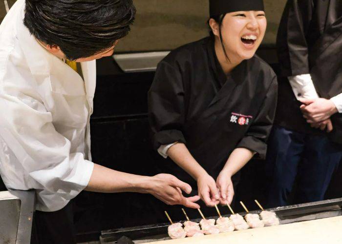 A guest laughing while making yakitori, guided by an expert.